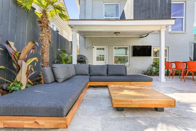 Sunny backyard patio with modern L-shaped wooden sectional topped with dark gray cushions, low wooden coffee table, palm and tropical plants, covered porch with wall-mounted TV and grill, and bright orange outdoor dining chairs.
