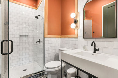 Modern bathroom with glass walk-in shower, white subway tile, black fixtures, terracotta accent wall, wood trim, white vanity and patterned terrazzo-style floor.