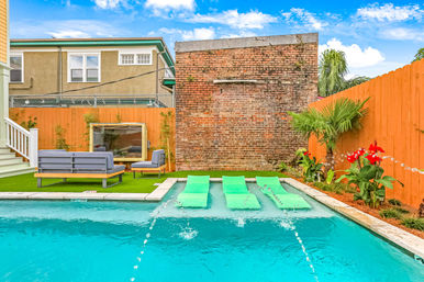 Urban backyard pool with three bright green floating loungers in turquoise water, fountain jets, artificial turf patio with gray outdoor sofa and chair, tall exposed brick wall, orange wooden fence, tropical plants and red flowers under a sunny blue sky.