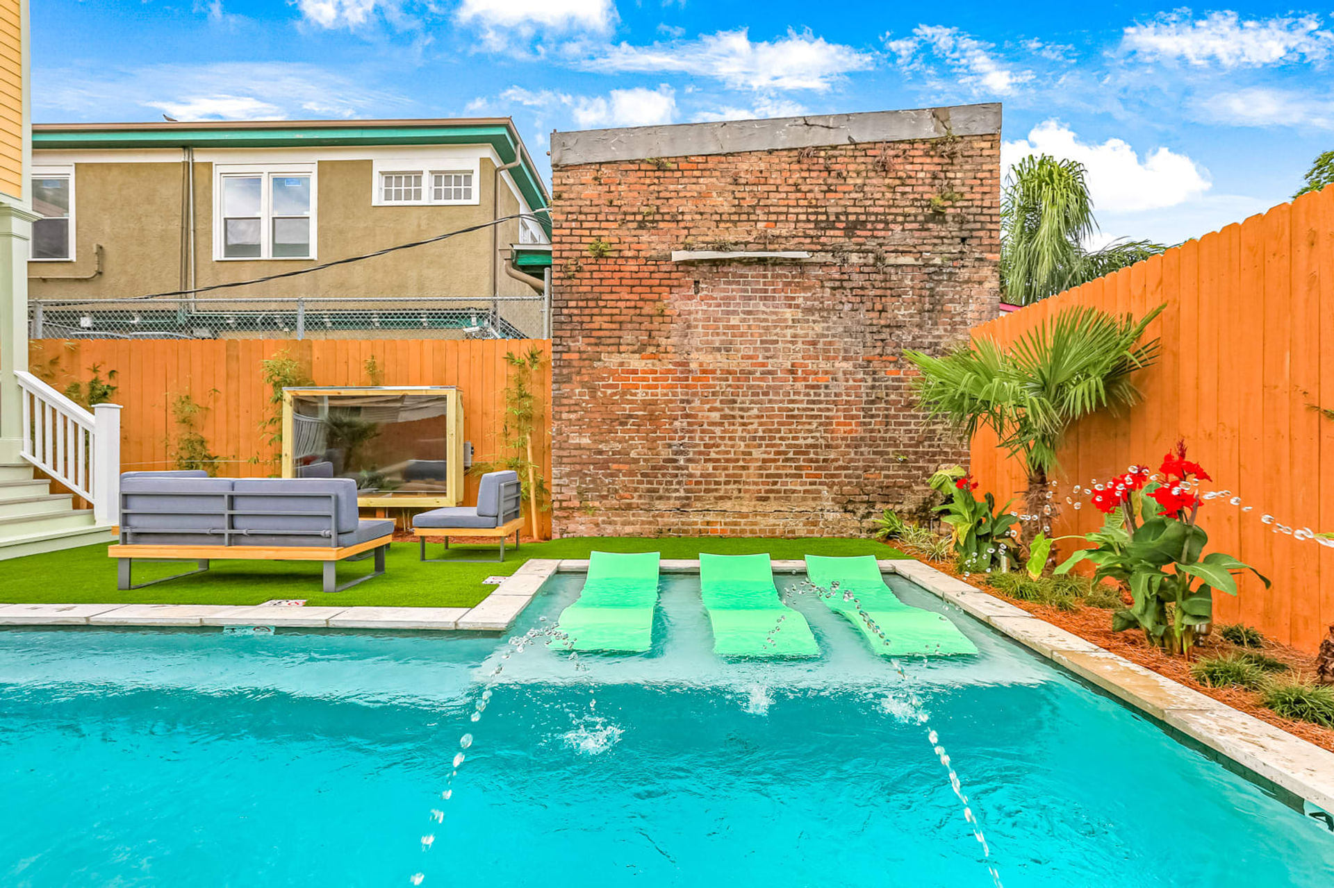 Urban backyard pool with three bright green floating loungers in turquoise water, fountain jets, artificial turf patio with gray outdoor sofa and chair, tall exposed brick wall, orange wooden fence, tropical plants and red flowers under a sunny blue sky.