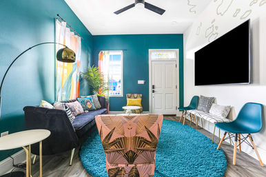 Vibrant contemporary living room and entryway with a teal accent wall, cozy navy sofa, patterned pink armchair, round turquoise rug, wall-mounted TV, colorful curtains and a potted plant.