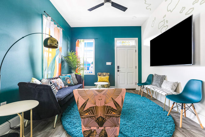 Vibrant contemporary living room and entryway with a teal accent wall, cozy navy sofa, patterned pink armchair, round turquoise rug, wall-mounted TV, colorful curtains and a potted plant.