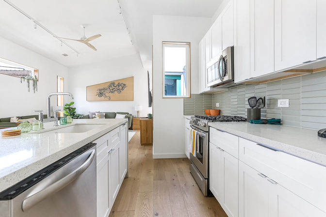 Bright, airy open-concept kitchen with white shaker cabinets, quartz island with sink, stainless steel appliances, green glass subway tile backsplash, and light wood floors opening to a sunlit living area