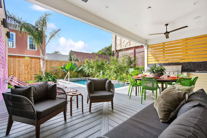 Covered modern backyard patio with gray lounge seating, green outdoor dining chairs and table, built-in grill, and a small rectangular pool surrounded by tropical plants and a wooden fence — bright, inviting outdoor living space.