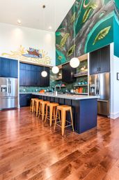 Vibrant modern open-concept kitchen with large island, six orange metal stools, globe pendant lights, teal subway tile backsplash, stainless appliances and a dramatic tree-and-leaf mural on a tall wall.
