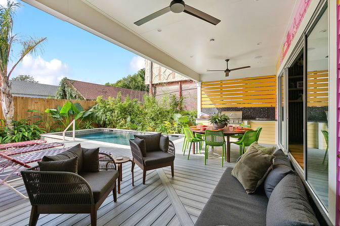 Sunny backyard patio oasis with covered deck and ceiling fans overlooking a rectangular plunge pool; grey wicker lounge seating, green dining chairs at a round table by a yellow slatted accent wall, tropical plants and modern outdoor grill