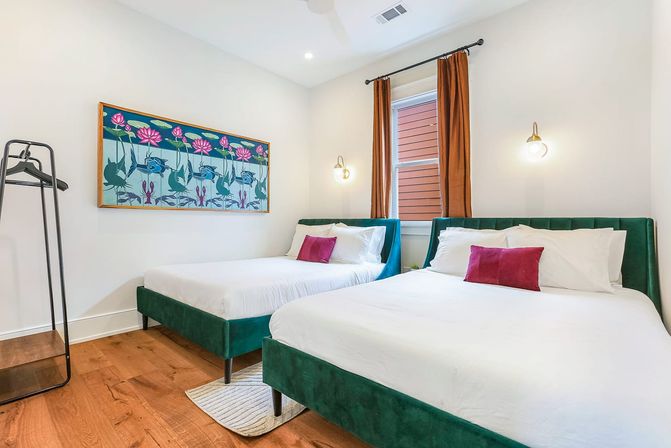 Bright modern guest bedroom in a city apartment with two green velvet beds dressed in white linens and magenta accent pillows, hardwood floors, colorful lotus wall art, rust-colored curtains, and wall-mounted pendant lights.