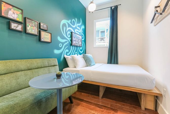 Cozy modern guest room with teal accent wall and swirling mural, white platform bed with pillows, green upholstered loveseat, round glass table, framed art, hardwood floors and a sunlit window