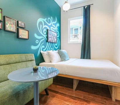 Cozy modern guest room with teal accent wall and swirling mural, white platform bed with pillows, green upholstered loveseat, round glass table, framed art, hardwood floors and a sunlit window