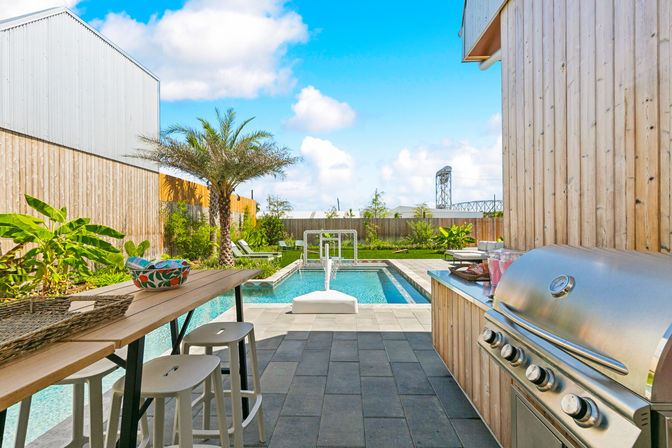 Sunny modern backyard with wood-clad patio, stainless-steel grill and bar seating beside a rectangular lap pool, palm tree and lounge chairs under a bright blue sky with a distant bridge visible