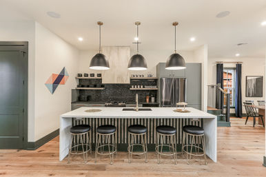 Modern open-concept kitchen with a large white island and five black stools, three black dome pendant lights, stainless refrigerator, speckled backsplash and light hardwood floors.