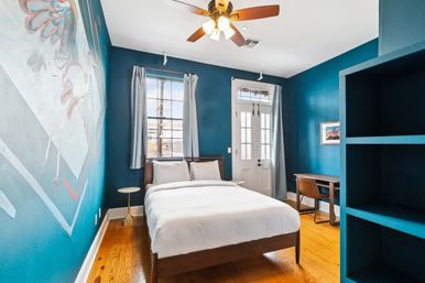 Cozy modern bedroom with deep teal accent walls, hardwood floors and ceiling fan, white-bedded queen bed, window and glass door with light curtains showing a palm-tree street view, small round side table and wooden desk with chair.