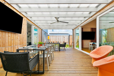Sunlit covered wooden patio and dining deck with a long table set for guests, ceiling fan, potted plants, wicker seating and two bright orange lounge chairs opening to a modern interior through sliding glass doors.