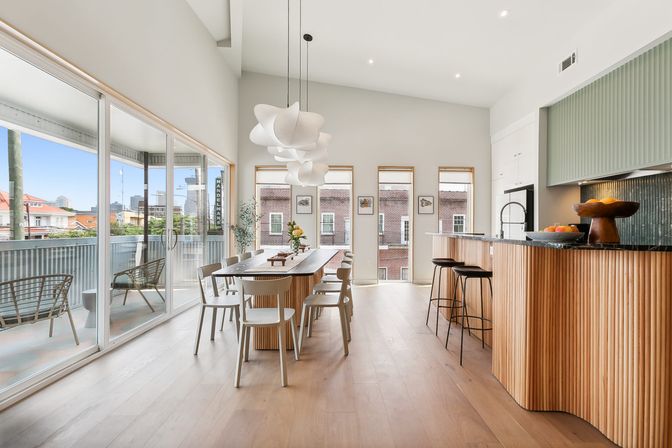 Bright modern open-plan dining and kitchen in an urban apartment — long wooden dining table, sculptural white pendant lights, curved wood-paneled breakfast island with bar stools, floor-to-ceiling sliding glass doors to a balcony and city skyline view.