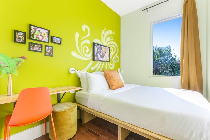 Cheerful lime-green bedroom with a white platform bed, orange accent pillow and chair, wooden desk, playful white wall mural and framed photos, palm-tree view through the window