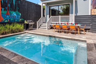 Modern backyard with rectangular turquoise swimming pool, paved stone deck, long outdoor dining table with orange chairs set for a meal, gas grill, colorful mural on a black fence, and gray house deck with sliding glass door.