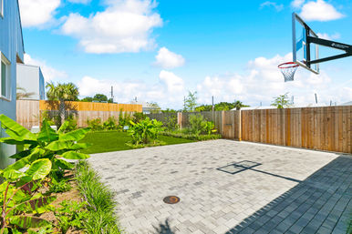 Sunny modern backyard with a paved half-court and basketball hoop, wooden privacy fence, manicured lawn and tropical landscaping under a bright blue sky.