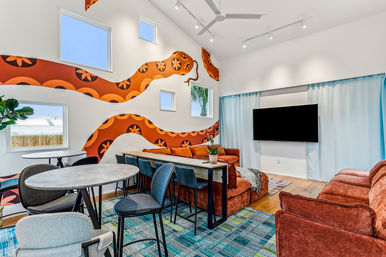 Bright contemporary lounge with high ceilings, bold orange snake mural on white walls, rust-colored sofas, wall-mounted TV with blue curtains, communal bar table with stools, round cafe tables, and a blue-green patterned rug under small square windows.