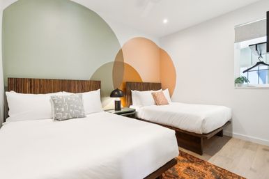 Bright modern guest room with two white-dressed beds and wooden slatted headboards, overlapping pastel circle mural on the wall, black bedside lamp on a small table, patterned rug and light wood floor.