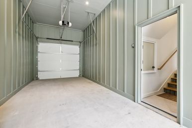 Bright, clean single-car attached garage with sage-green vertical paneled walls, concrete floor, automatic overhead door opener, and interior door leading to home stairs.