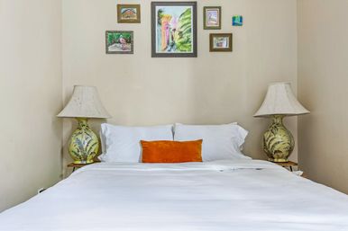 Cozy bedroom with crisp white bedding and an orange lumbar pillow, flanked by matching palm-motif table lamps and a small gallery wall of colorful framed paintings on a neutral wall.