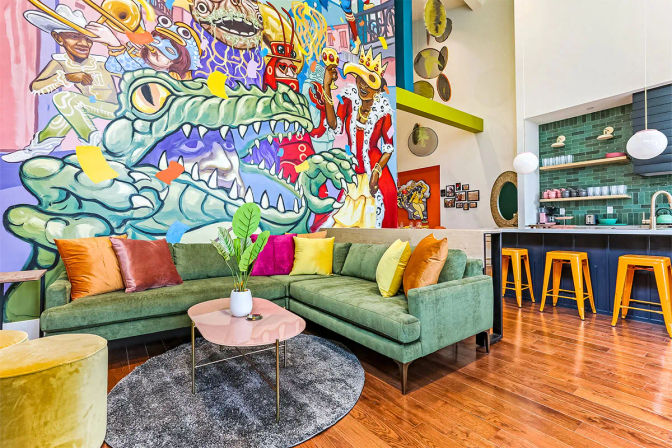 Vibrant urban lounge featuring a bold cartoon crocodile mural, green velvet L-shaped sofa with colorful throw pillows, pink coffee table on a round gray rug, hardwood floors and an open kitchen bar with green subway tile backsplash and yellow stools.
