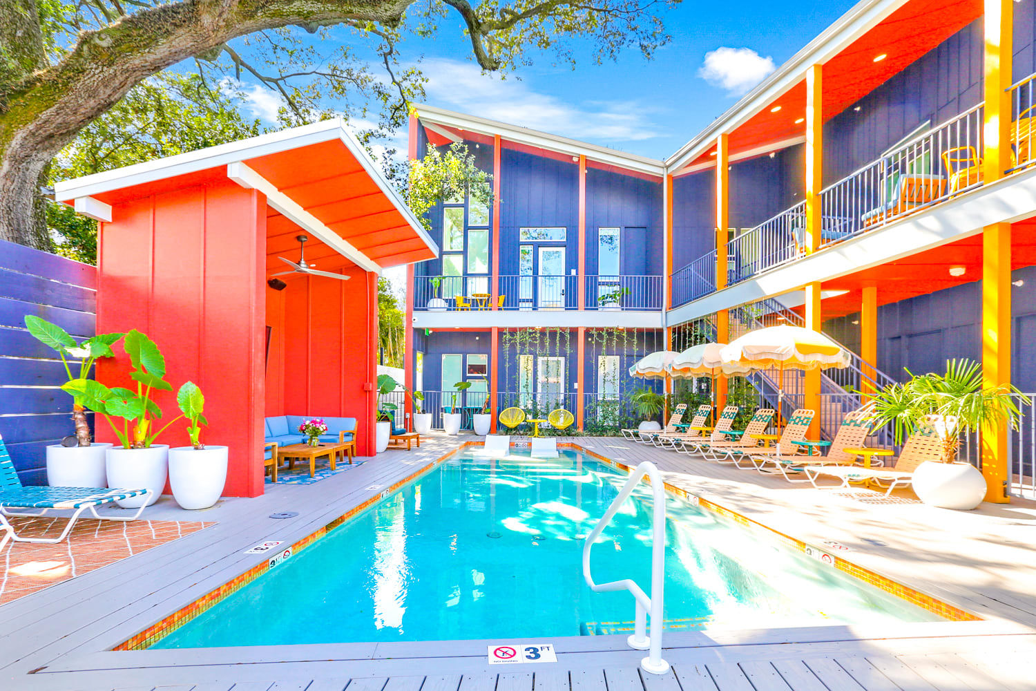 Vibrant outdoor courtyard pool at a colorful boutique hotel with a red cabana, loungers and umbrellas, two-story balcony, tropical plants and bright blue sky.