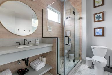 Spa-like modern bathroom with glass-enclosed corner shower, beige subway tile, floating vanity with matte-black faucet and round mirror, white toilet and framed wall art