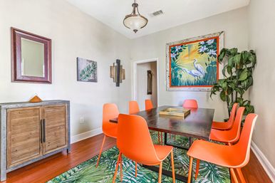 Contemporary dining room interior with six bright orange chairs around a dark wood table on a green tropical-leaf rug, colorful heron wall art, wooden sideboard, ceiling light, and tall potted plant.