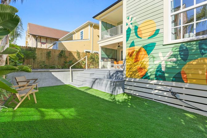 Sunny residential backyard patio with vibrant citrus mural on house siding, bright artificial turf, gray deck steps, wooden lounge chairs, tropical plants and a second-story balcony.