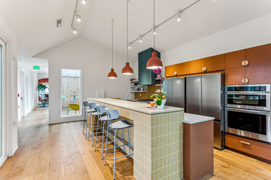Bright modern open-plan communal kitchen in a co‑living space with a long tiled island and metal barstools, terracotta pendant lights, stainless steel appliances, wood floors, and a potted orchid on the counter.
