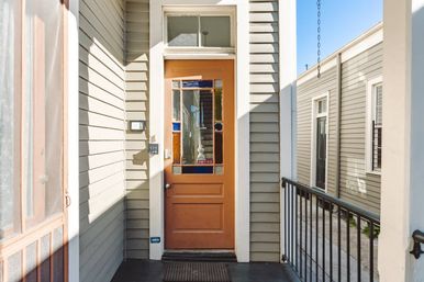 Sunlit urban front porch with an orange wooden door featuring stained-glass panels, keypad entry and doormat, flanked by gray clapboard siding and a black metal railing.