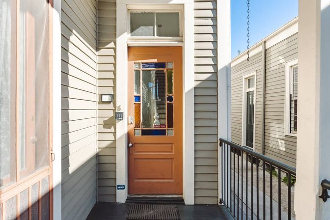 Sunlit urban front porch with an orange wooden door featuring stained-glass panels, keypad entry and doormat, flanked by gray clapboard siding and a black metal railing.
