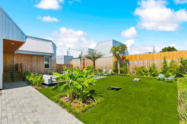 Sunny modern backyard with green lawn and palm trees, paved walkway to a wood-and-metal house, patio lounge seating, sun loungers by a small pool, and cornhole boards for outdoor fun.