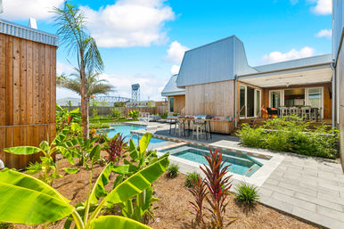 Sunny modern wood‑clad backyard with swimming pool and built‑in spa, tropical landscaping with palms and banana plants, outdoor dining and bar seating on a deck with a bridge visible in the distance