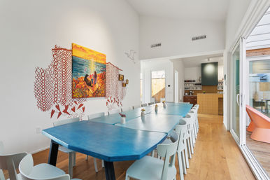 Modern coastal-inspired dining room with a long blue communal table and white chairs, beach painting with coral wall art, hardwood floors, sliding glass doors to a patio, and an open kitchen in the background
