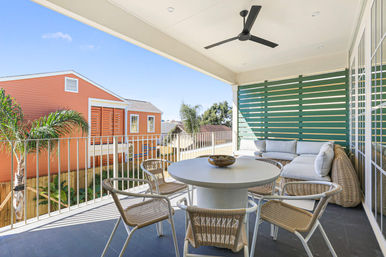 Sunny second-floor balcony with a round dining table, woven chairs, cushioned wicker sectional, ceiling fan and green slatted privacy screen, overlooking palm trees and colorful beach-style houses.