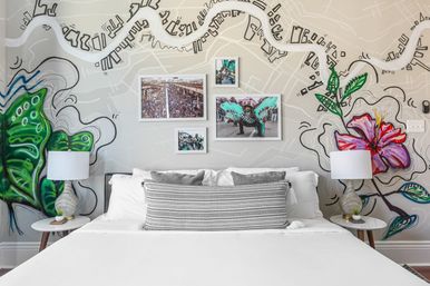 Modern boutique bedroom with white bed and patterned lumbar pillow, bedside lamps, gallery wall of carnival photos and a colorful map-style mural with green butterfly and pink hibiscus.
