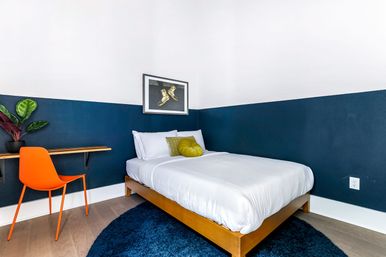 Bright modern bedroom with wooden platform bed in white linens against a navy half-wall, round navy rug, orange desk chair at a floating wood desk, green accent pillow and framed artwork.