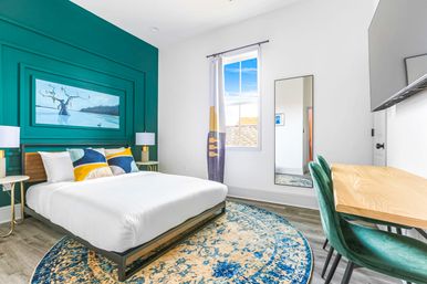 Bright modern coastal-inspired bedroom with teal accent wall and seascape painting, white bed with colorful pillows, round blue-beige rug, wood desk with green velvet chairs, wall-mounted TV and sunny window.
