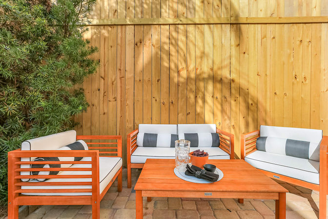 Sunlit backyard patio with contemporary orange wooden outdoor seating and white cushions, matching coffee table on pavers by a wooden privacy fence and leafy shrub.