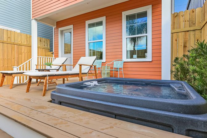Sunny backyard deck with bubbling hot tub, two wooden lounge chairs and a turquoise bistro set in front of an orange clapboard house and wooden privacy fence, palm tree reflected in the window.