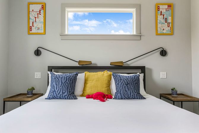 Modern coastal-inspired bedroom with crisp white bedding, two blue patterned pillows and a yellow accent pillow, a small red plush lobster on the bed, a high window showing blue sky, black swing-arm wall lamps and wooden nightstands with potted succulents.