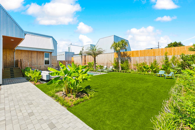 Sunny modern backyard with lush green lawn, tropical banana plants and palm trees, wooden fence, metal-clad contemporary house, patio seating, sun loungers and a small pool under a bright blue sky.