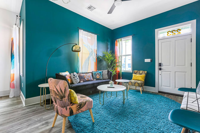 Vibrant teal-walled living room with colorful mid-century modern chairs and sofa, patterned pillows, round white coffee tables on a blue shag rug, arched brass floor lamp, potted palm and white entry door
