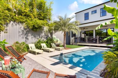 Sunny backyard pool at a modern two-story home with a palm tree, tropical landscaping, green chaise lounges, wooden lounge chairs and a drink on a side table.