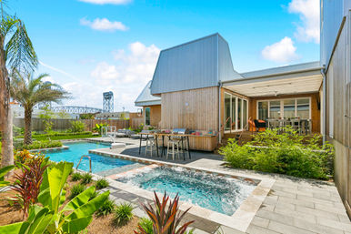 Sunny resort-style backyard with a rectangular pool and bubbling spa beside a modern wood-clad home, palm trees, deck seating and dining area, and an industrial bridge visible beyond the fenced yard.