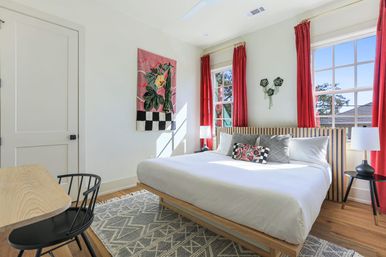 Sunlit modern bedroom with white bedding on a wooden slatted bed, red curtains framing two windows, floral wall tapestry, patterned rug, bedside lamps and a small desk with black chair.