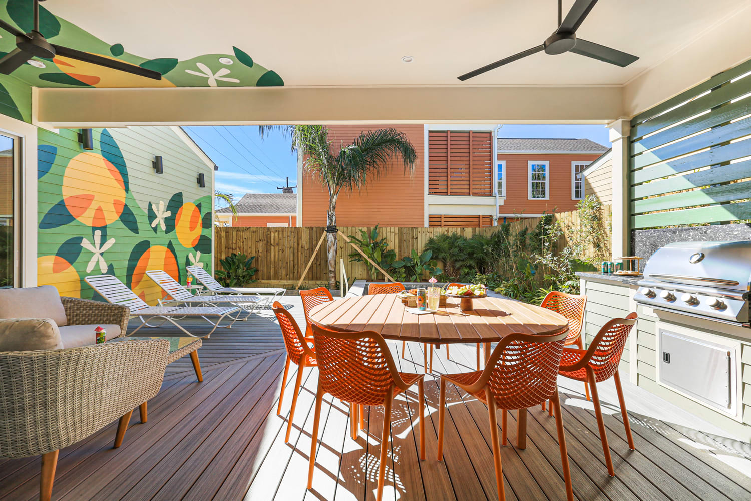 Sunny backyard deck with round wooden dining table and orange chairs, stainless grill, lounge chairs, palm tree and colorful citrus mural