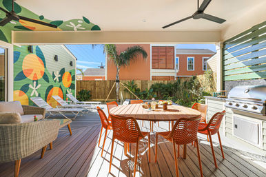 Sunny backyard deck with round wooden dining table and orange chairs, stainless grill, lounge chairs, palm tree and colorful citrus mural